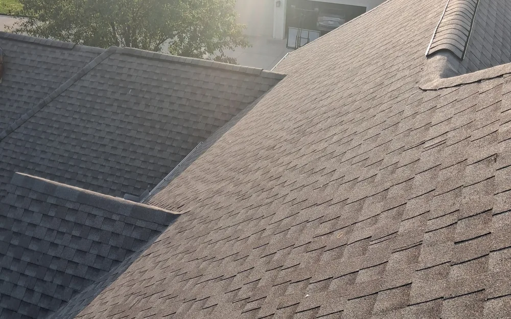 Aerial view of a completed shingle roof with multiple ridge lines and slopes, photographed in bright sunlight with a tree and neighboring garage in the background.
