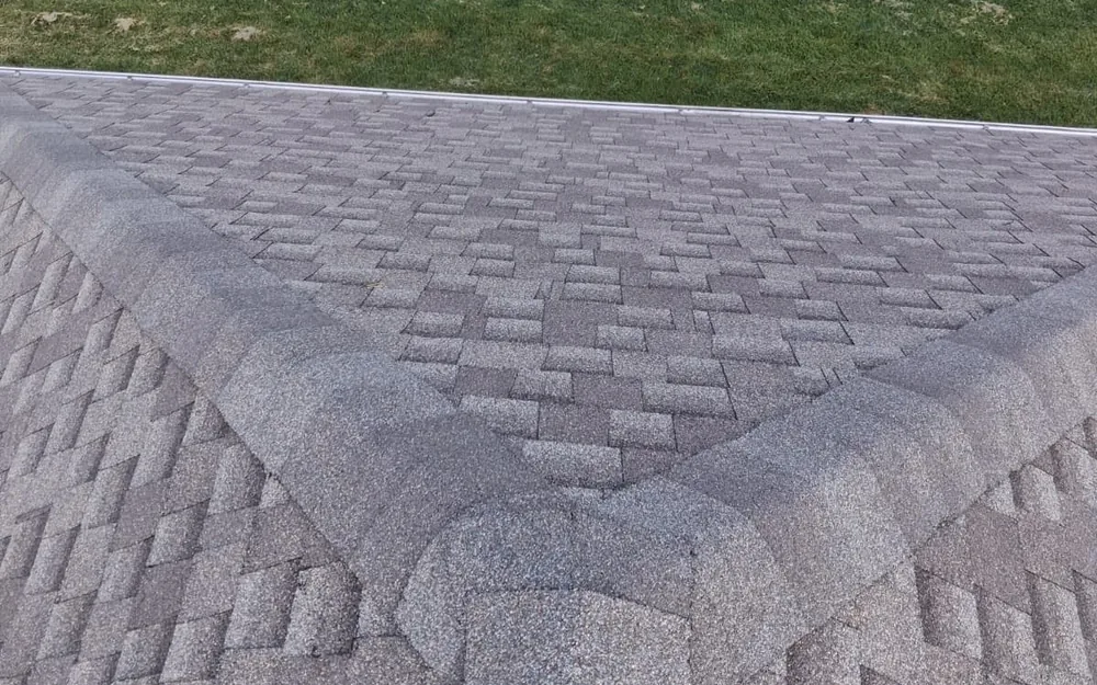 Close-up of a finished shingle roof hip and valley intersection showing clean, uniform shingle alignment, with a grass lawn visible at the eave.