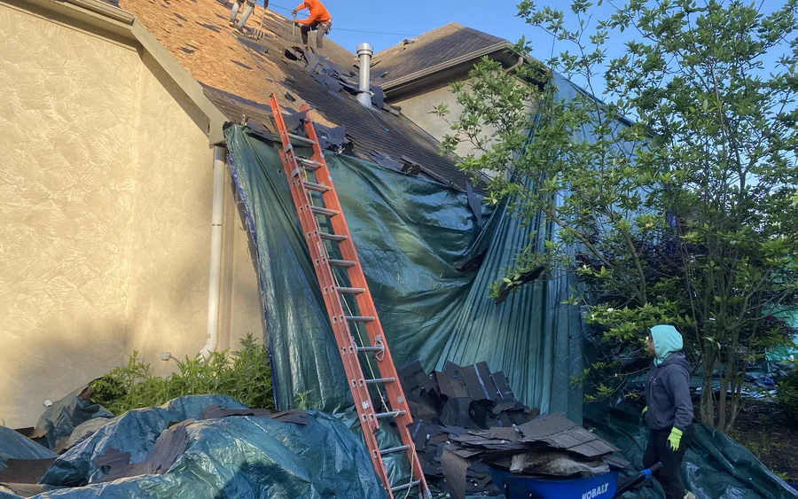 Roofing crew securing a heavy-duty tarp over a home's siding and landscaping during a roof tear-off, with a ladder and torn shingle debris staged below.