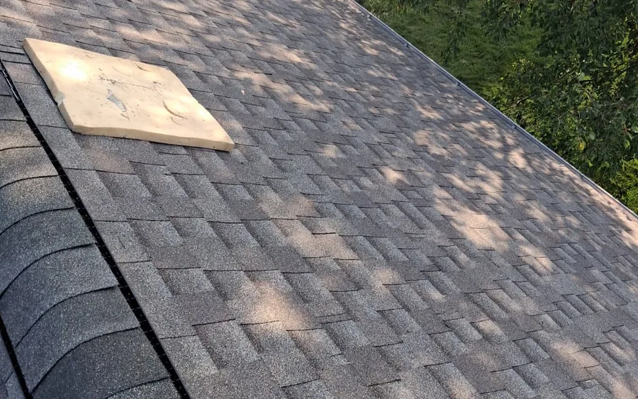 Close-up of a finished shingle roof and ridge line, with a spare shingle piece resting on the surface and dappled tree shade across the slope.