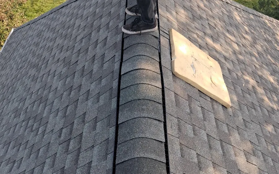 Roofer's legs and boots visible while installing ridge cap shingles along a roof peak, with a folded shingle bundle staged nearby and a lawn visible below.