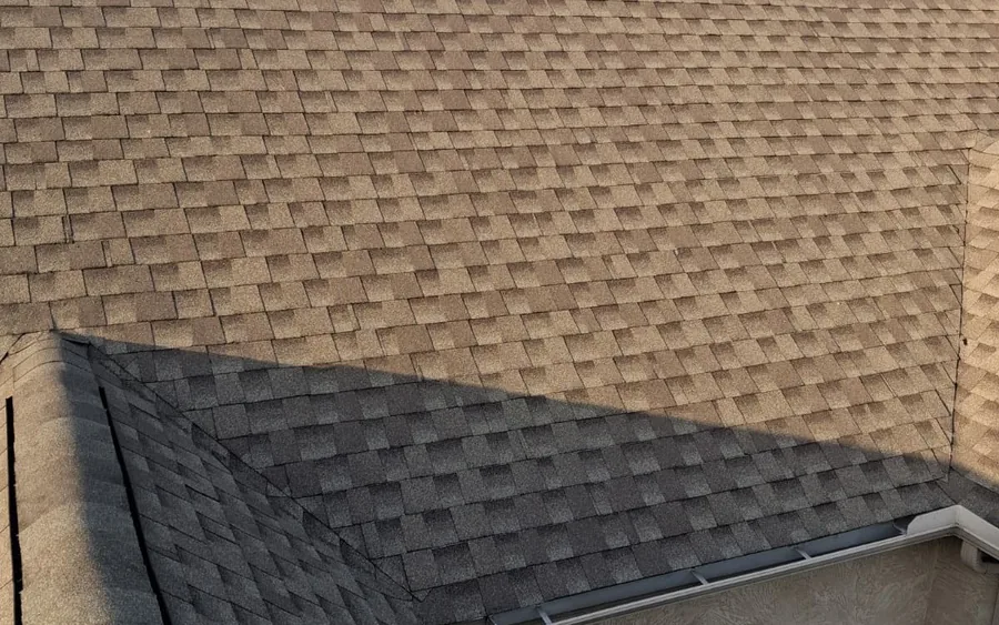 Aerial view of a completed asphalt shingle roof showing two adjoining roof planes meeting at a hip, with a small gable dormer, photographed in afternoon light.