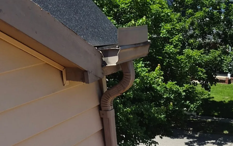 Close-up looking up at a roof eave where the shingle edge, gutter, downspout, and vinyl siding trim meet at a gable corner.