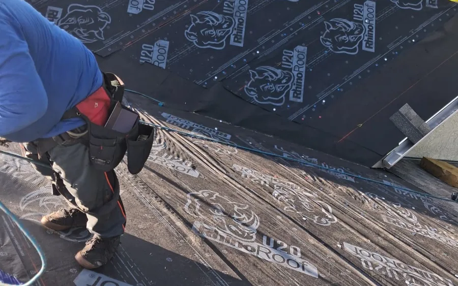 Roofer installing synthetic underlayment on a roof deck, with a tool belt, tree line, and torn-off roofing material visible.