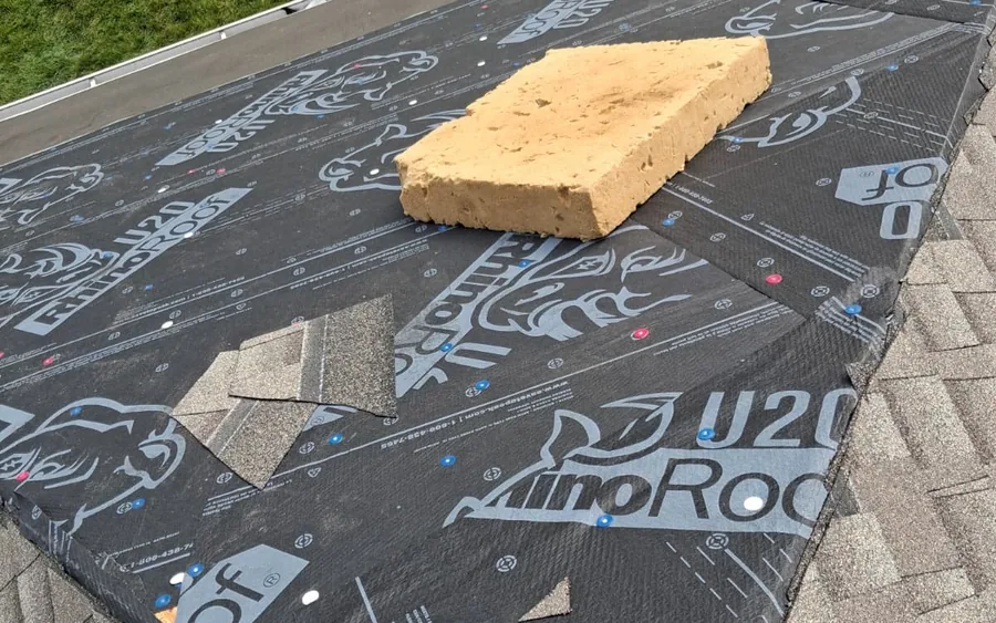 Synthetic underlayment installed over a roof deck, with a removed ridge cap shingle and torn shingle scraps staged on top before cleanup.